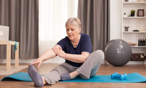 Active old woman exercising on the yoga mat in her comfortable apartment. Old person pensioner online internet exercise training at home sport activity with dumbbell, resistance band, swiss ball at elderly retirement age